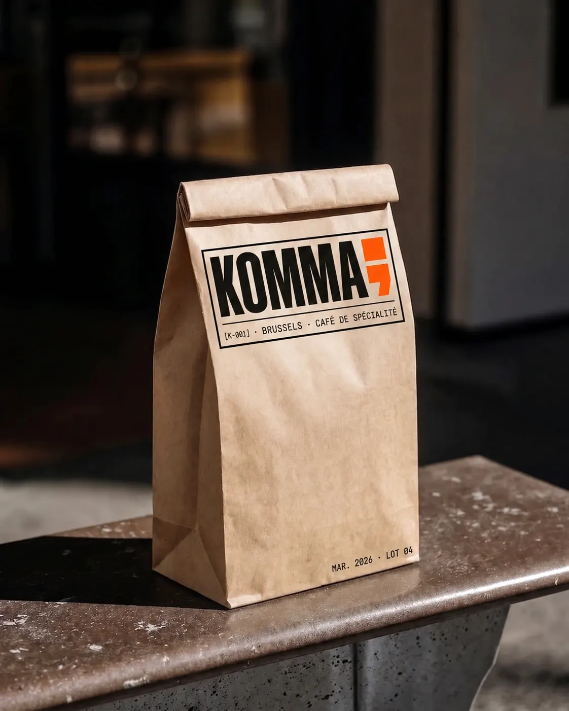 Sac à emporter en kraft brut KOMMA, posé sur un comptoir béton, avec logotype et code [K-001] visible.